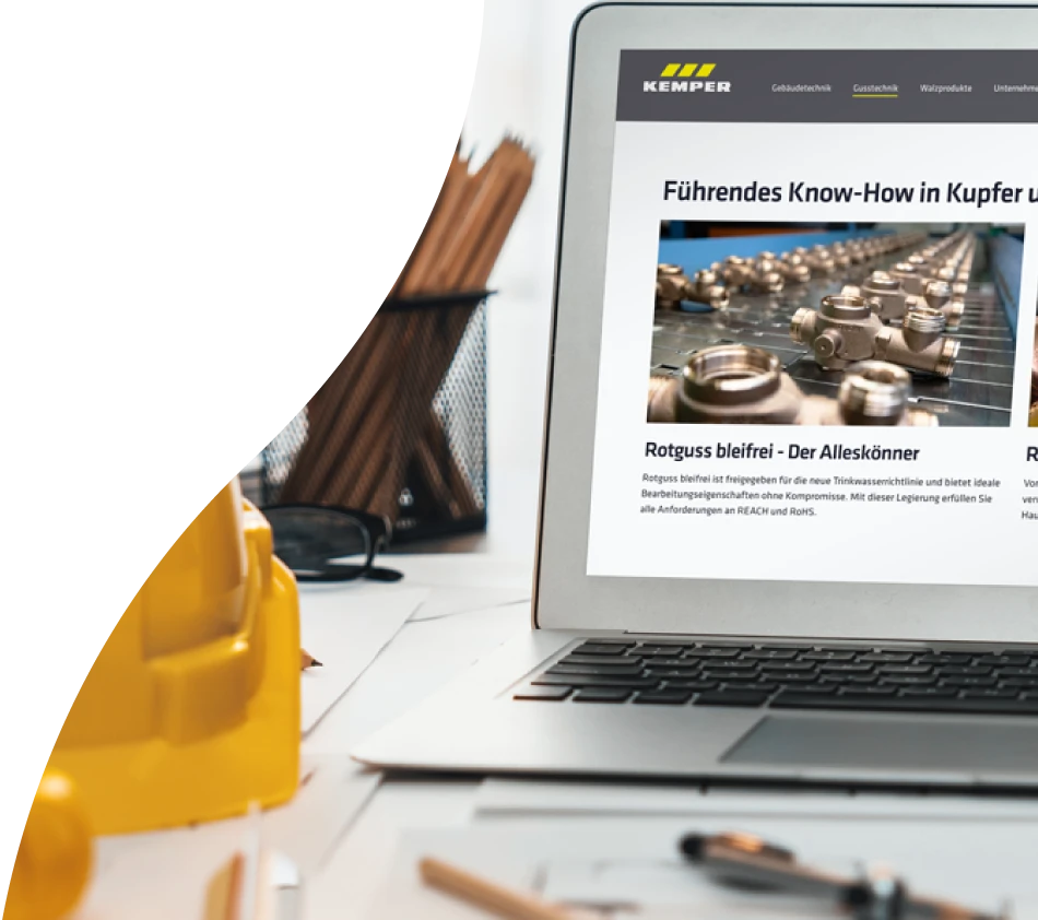 Ein aufgeklappter Laptop auf einem Tisch zeig den Screen des Onlineshops von KEMPER, der mit crossbase umgesetzt wurde. Auf dem Tisch befinden sich eingerollte Pläne, ein Schutzhelm für Baustellen, eine Pflanze, eine Brille und ein Stiftebecher.